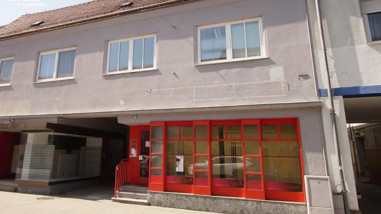 Gebäudefassade mit grauer Putzfassade und roten Schaufenstern im Erdgeschoss.
