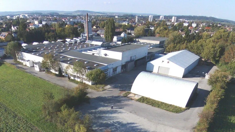 Luftaufnahme eines Industriegebiets mit mehreren Hallen, umliegender Grünfläche und städtischem Hintergrund.