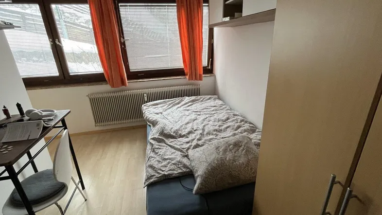 Helles Schlafzimmer mit Einzelbett, Schreibtisch und großem Fenster mit Jalousien.