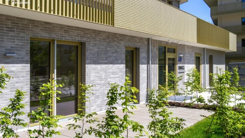 Moderne Erdgeschosswohnung mit gepflasterter Terrasse und junger Bepflanzung vor einer hellgrauen Ziegelfassade.
