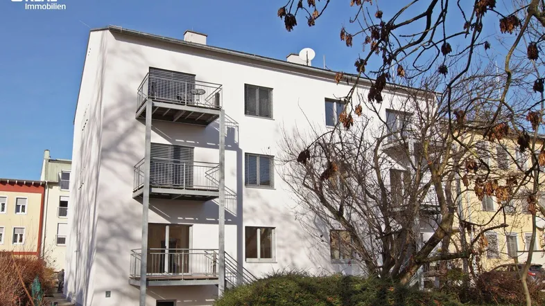 Weißes Mehrfamilienhaus mit mehreren Balkonen und Bäumen im Vordergrund unter blauem Himmel.