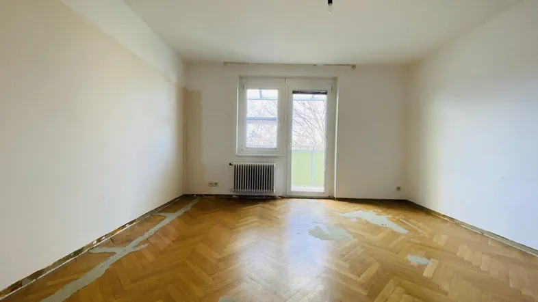 Heller Raum mit Fischgrätparkett, Heizkörper und Zugang zu einem Balkon, der eine Renovierung benötigt.