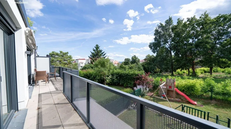 Großer Balkon mit Metallgeländer, Sitzgelegenheiten und Blick auf einen Spielplatz und viel Grün.