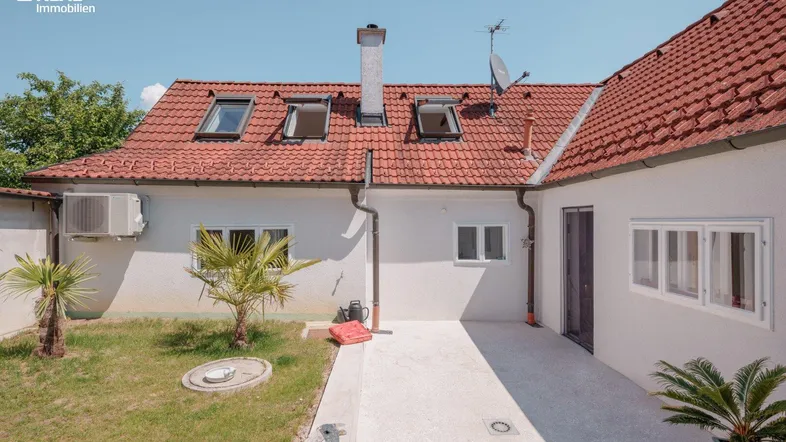 Hausrückseite mit kleiner Rasenfläche, gepflasterter Terrasse und Dachfenstern unter blauem Himmel.