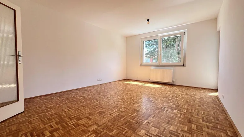Helles Zimmer mit Fischgrätparkettboden, weißen Wänden und Fenster, das viel Tageslicht hereinlässt.