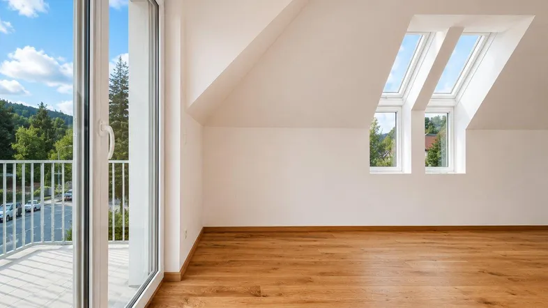 Helles Zimmer im Dachgeschoss mit Holzboden, Schrägdecken und Zugang zu einem Balkon mit Blick ins Grüne.