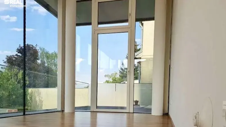 Heller Raum mit großen Glasfronten, Holzboden und Zugang zu einem Außenbereich.