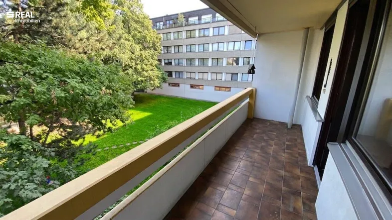 Balkon mit braunen Fliesen und Blick auf eine grüne Rasenfläche und Bäume.