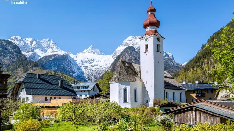 Idyllisches Dorf mit traditioneller Kirche und schneebedeckten Bergen im Hintergrund unter blauem Himmel.