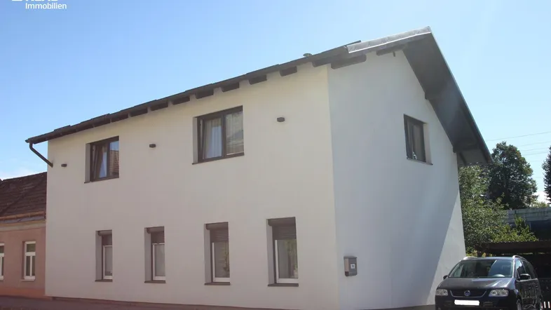 Weiß verputztes Einfamilienhaus mit Satteldach und dunklen Fenstern unter blauem Himmel.