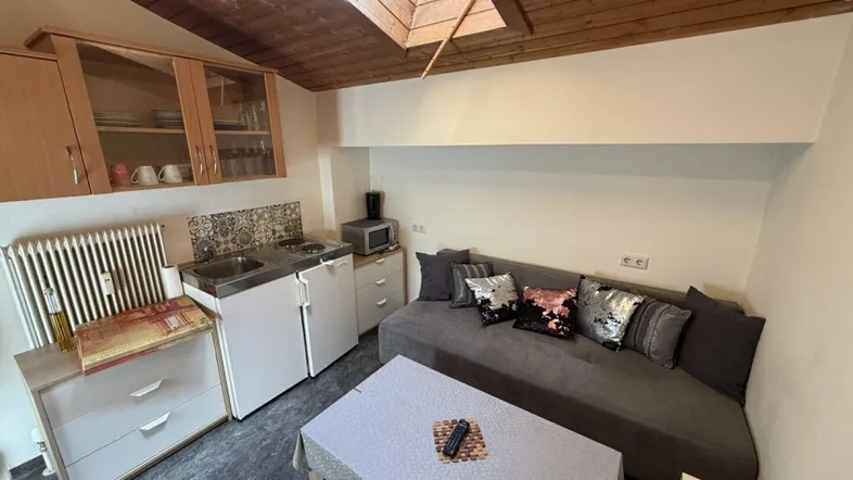 Helles Wohnzimmer mit integrierter Küchenzeile, Dachschräge und Holzbalkendecke.