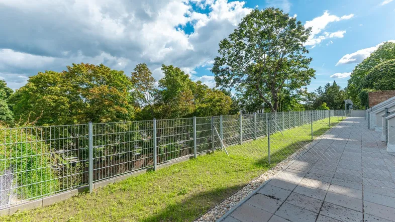 Außenbereich mit gepflastertem Weg, Grünfläche und Metallzaun unter bewölktem Himmel.