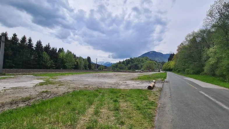 Ungenutztes Grundstück neben einer Straße und Bahngleisen mit Bergkulisse im Hintergrund.