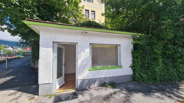Kleines weißes Gebäude mit grünen Akzenten und üppiger Vegetation an der Fassade.