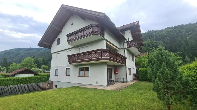 Mehrstöckiges Wohnhaus mit mehreren Balkonen und Satteldach, umgeben von Grünflächen und Bergen.