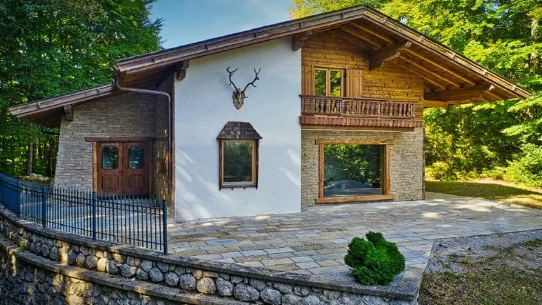 Charmantes Landhaus mit Stein- und Holzfassade, kleinem Balkon und gepflasterter Terrasse, umgeben von Bäumen.