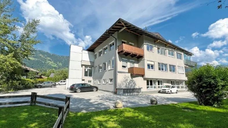 Modernes Gebäude mit mehreren Etagen, Balkonen und Parkplätzen, umgeben von grüner Landschaft und Bergen.