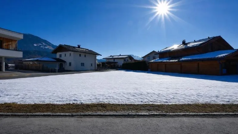 Sonniges Grundstück mit Schnee, umgeben von Häusern und einer beeindruckenden Bergkulisse.