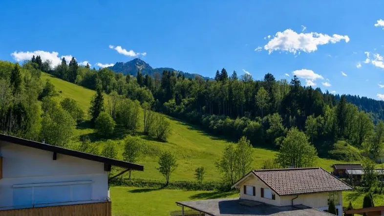 Panoramablick auf grüne Hügel und bewaldete Berge mit traditionellen Häusern unter blauem Himmel.