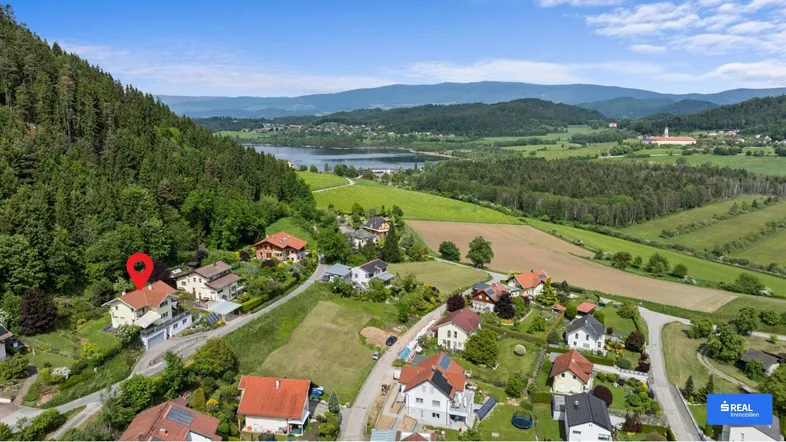 Luftaufnahme der Wohnsiedlung mit umliegender Natur, Wald, See und Bergen im Hintergrund.
