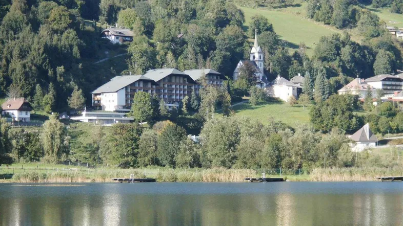 Panoramablick auf den See mit Gebäuden, Kirche und bewaldeten Bergen im Hintergrund.