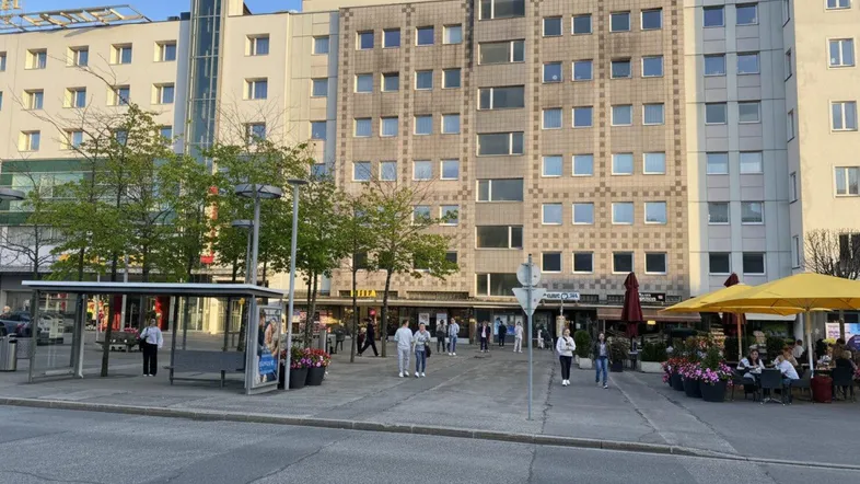 Blick auf eine belebte Straße mit modernen Gebäuden, Geschäften und einem Café im Freien unter blauem Himmel.
