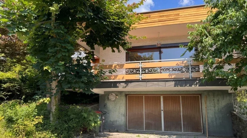 Hausfassade mit Balkon, integrierter Doppelgarage und üppiger Vegetation im Vordergrund.