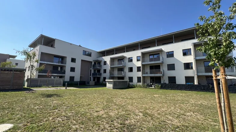 Modernes Mehrfamilienhaus mit heller Fassade und Grünfläche im Vordergrund unter blauem Himmel.