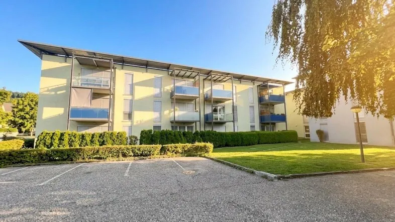 Modernes Mehrfamilienhaus mit Balkonen, umgeben von Grünflächen und einem Parkplatz unter blauem Himmel.