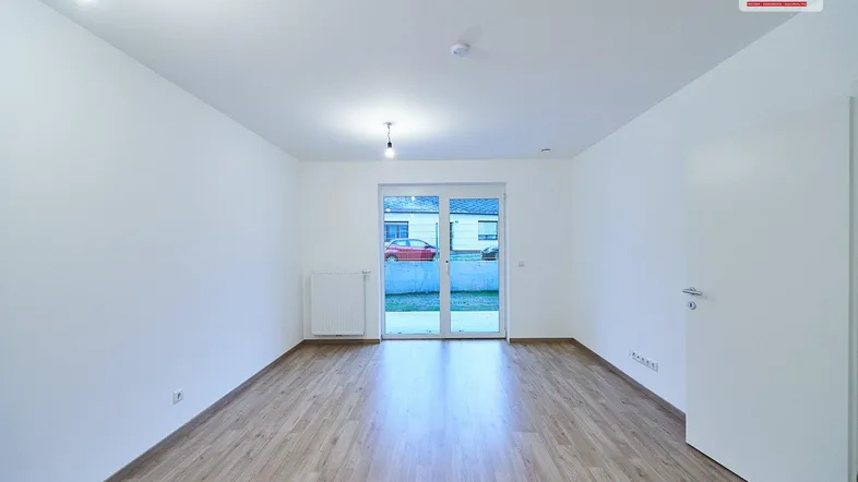 Helles, leeres Zimmer mit Laminatboden und Zugang zu einem Außenbereich durch eine Glastür.