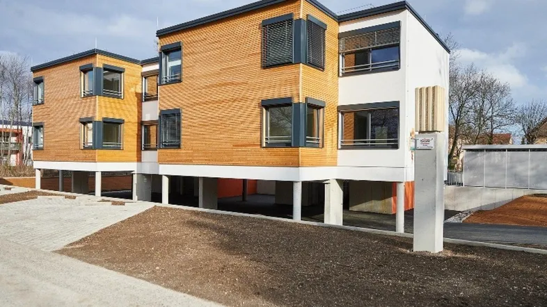 Modernes Mehrfamilienhaus mit Holzfassade und großen Fenstern, umgeben von Grünflächen und Parkplätzen.
