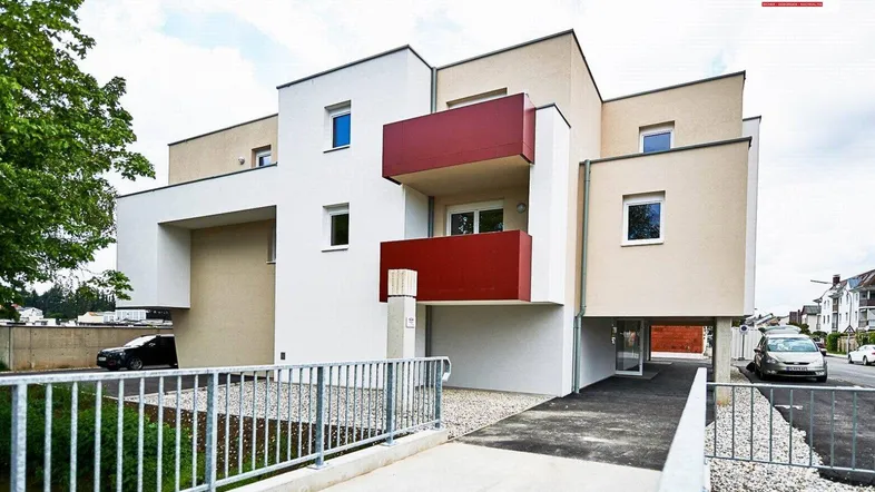 Modernes Mehrfamilienhaus mit heller Fassade, roten Balkonen und gepflegter Außenanlage.