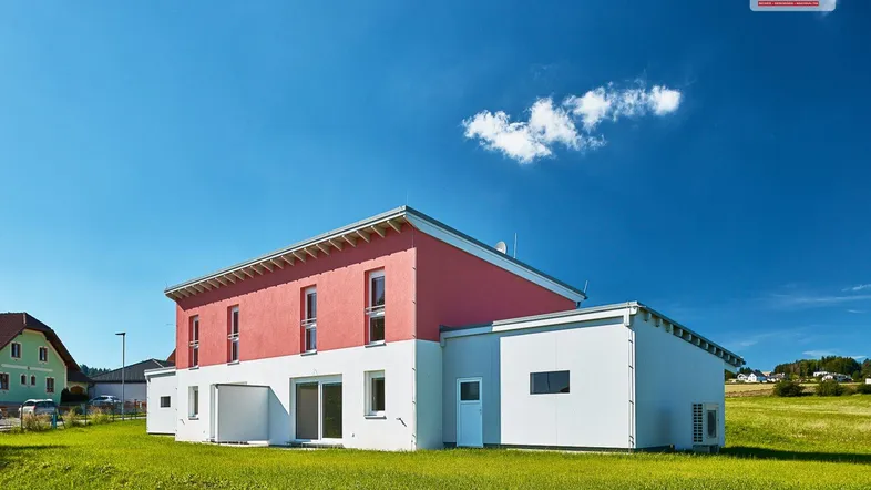 Modernes Einfamilienhaus mit roter Fassade und weißem Sockel auf grünem Rasen unter blauem Himmel.