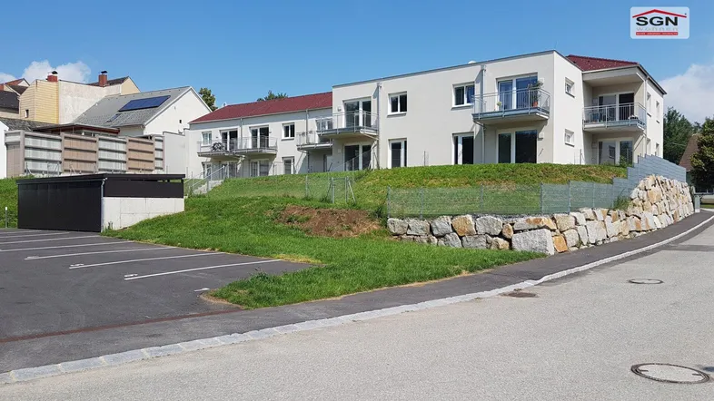 Modernes Mehrfamilienhaus mit Balkonen, Parkplätzen und einer Natursteinmauer in sonniger Lage.