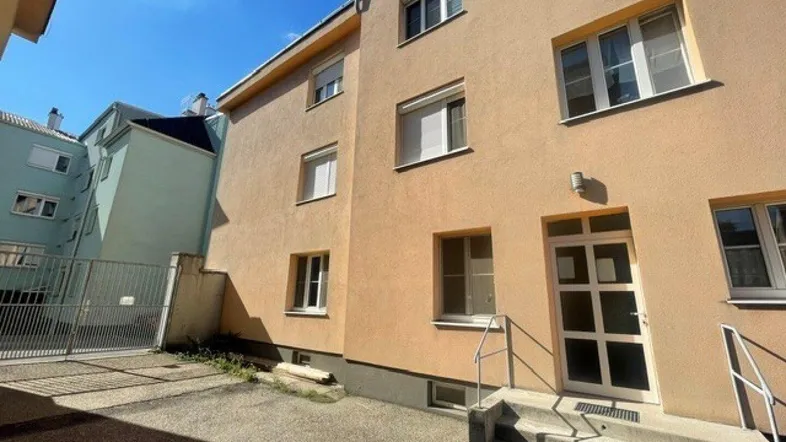 Fassade eines Mehrfamilienhauses mit Eingangsbereich und gepflastertem Vorplatz unter blauem Himmel.