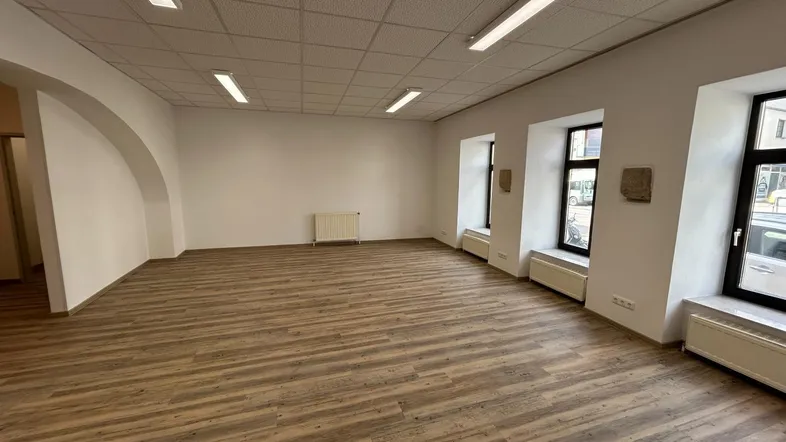Geräumiger, leerer Gewerberaum mit Laminatboden, abgehängter Decke und mehreren Fenstern zur Straße.