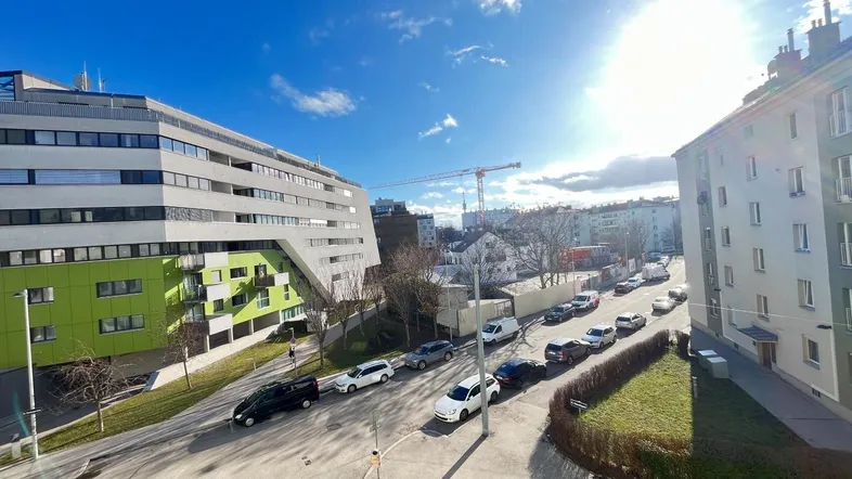 Sonnige Straßenansicht mit modernen und traditionellen Gebäuden sowie geparkten Autos unter blauem Himmel.