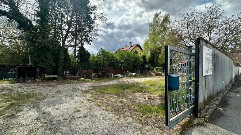 Blick auf ein großes Grundstück mit Schotterweg, Bäumen und einem Haus im Hintergrund unter bewölktem Himmel.
