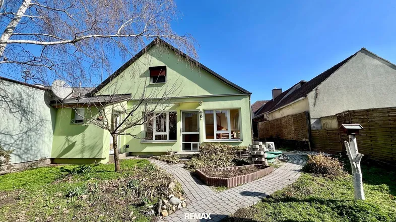 Grünes Einfamilienhaus mit Satteldach und gepflegtem Vorgarten unter blauem Himmel.