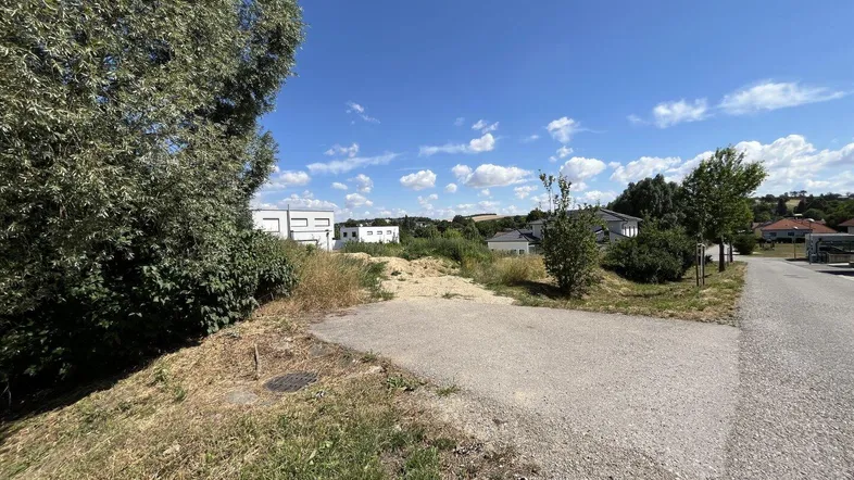 Ungenutztes Grundstück mit angrenzender Straße und Blick auf umliegende Häuser unter blauem Himmel.