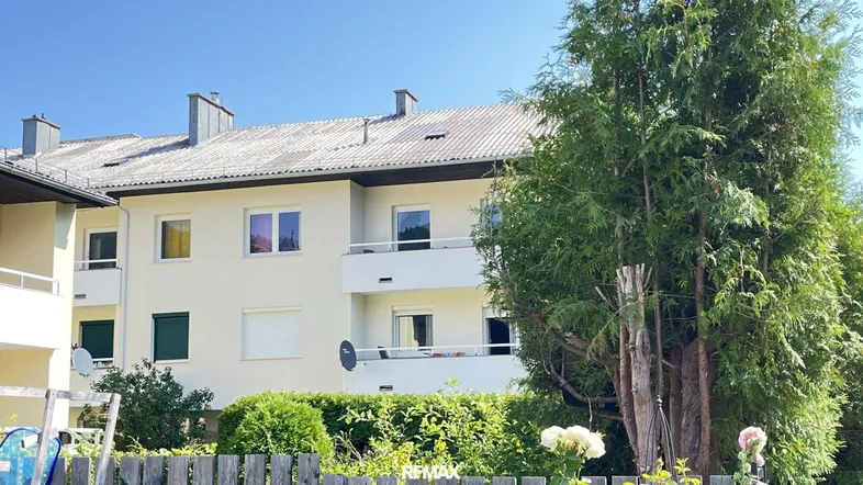 Mehrfamilienhaus mit heller Fassade, Balkonen und Satteldach umgeben von Bäumen.