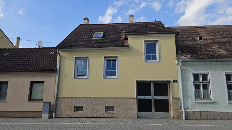 Gelbe Fassade eines Wohnhauses mit Satteldach und Fenstern zur Straße hin unter blauem Himmel.
