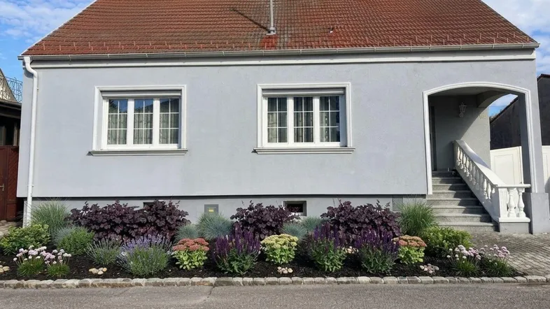 Hausfassade mit neu angelegtem Vorgarten und dekorativer Bepflanzung unter blauem Himmel.
