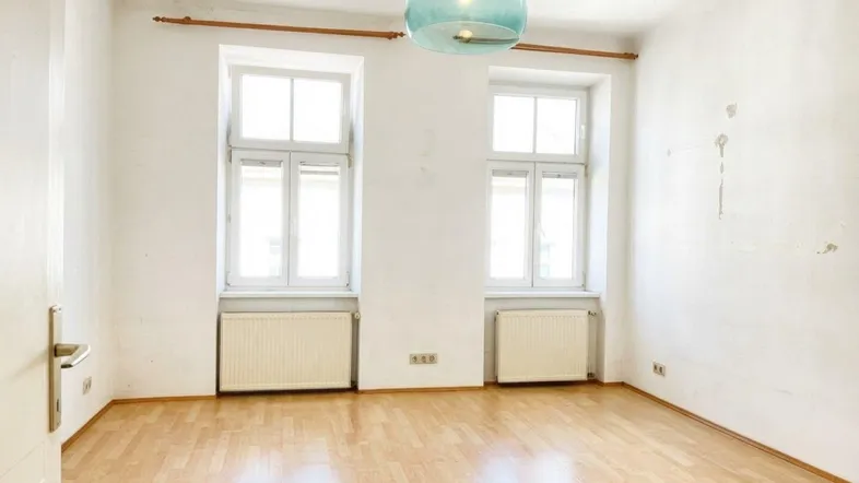 Helles, leeres Zimmer mit Parkettboden und zwei großen Fenstern, bereit zur individuellen Gestaltung.