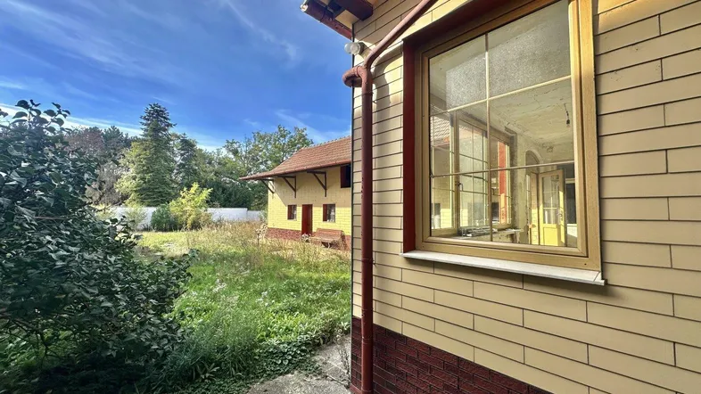Seitenansicht eines Hauses mit gelber Fassade, roten Ziegeln und einem verwilderten Gartenbereich.