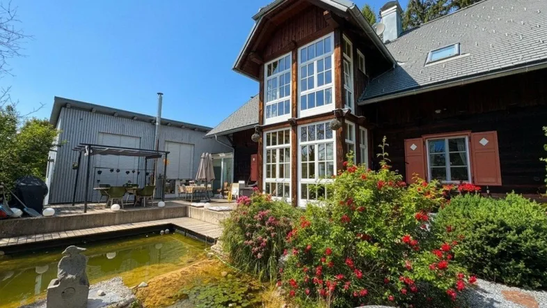 Charmantes Holzhaus mit großzügigem Gartenbereich, Teich und Terrasse unter blauem Himmel.