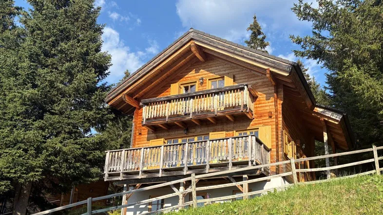 Charmantes Holzchalet in alpiner Umgebung mit zwei Balkonen und umgebenden Nadelbäumen unter blauem Himmel.