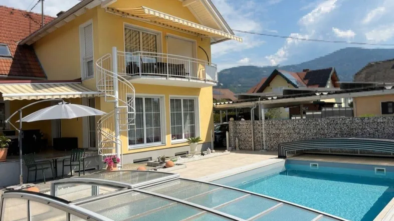 Gelbes Haus mit Swimmingpool, überdachter Terrasse und Balkon, umgeben von Bergen.