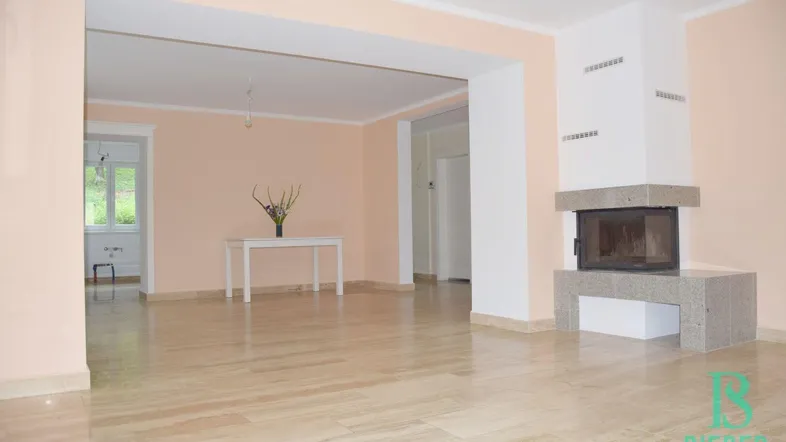 Geräumiges Wohnzimmer mit hellem Fliesenboden, Kamin und Zugang zu weiteren Räumen.