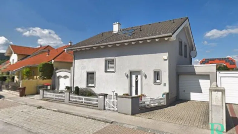 Graues Einfamilienhaus mit Vorgarten, Garage und gepflasterter Einfahrt unter blauem Himmel.